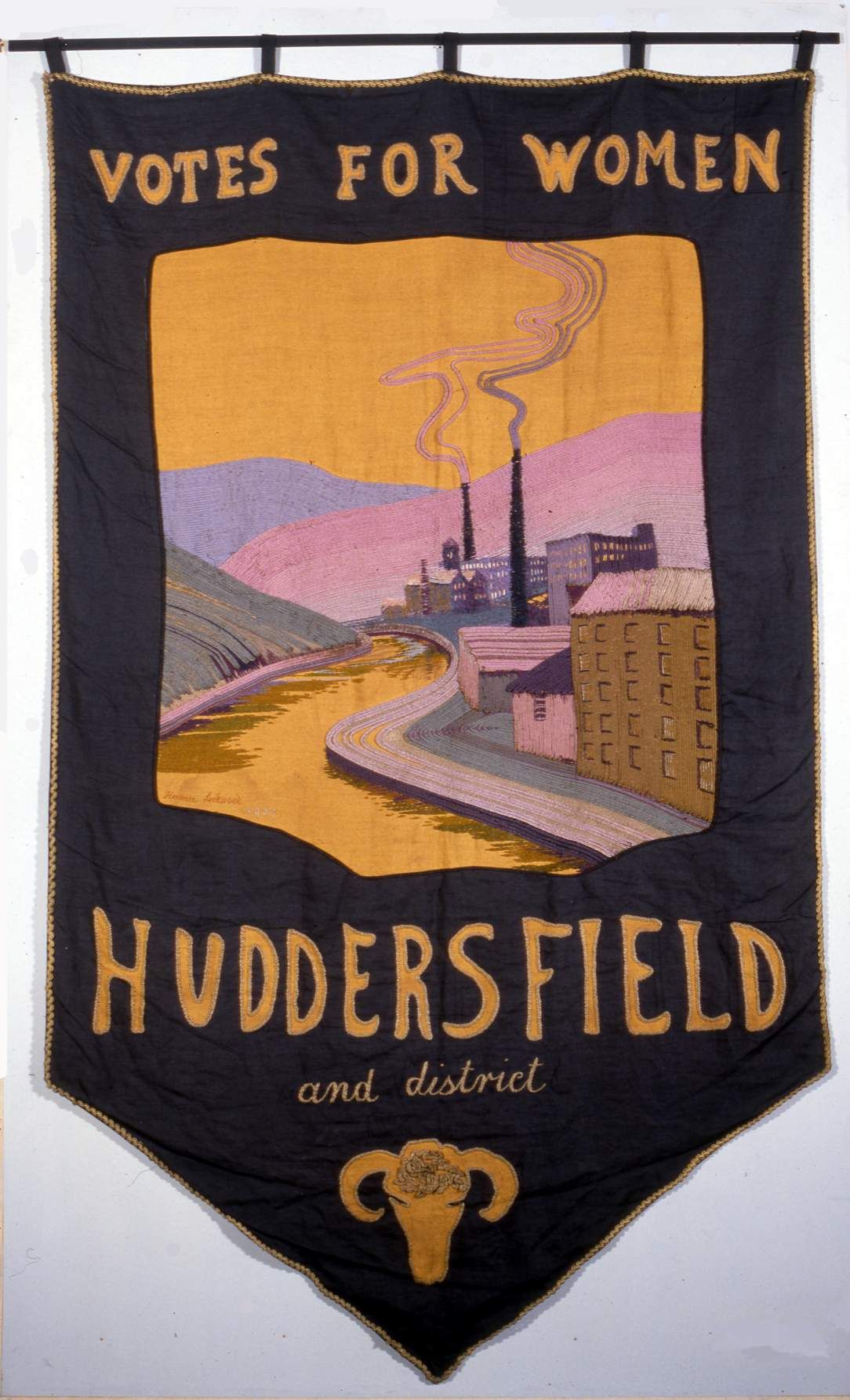 A dark fabric banner with yellow text reading "Votes for Women" in capitals at the top and "Huddersfield and district" at the bottom. The centre shows a colourful illustration of a river flowing through an industrial town with factories and smokestacks. A yellow ram’s head emblem is at the bottom.