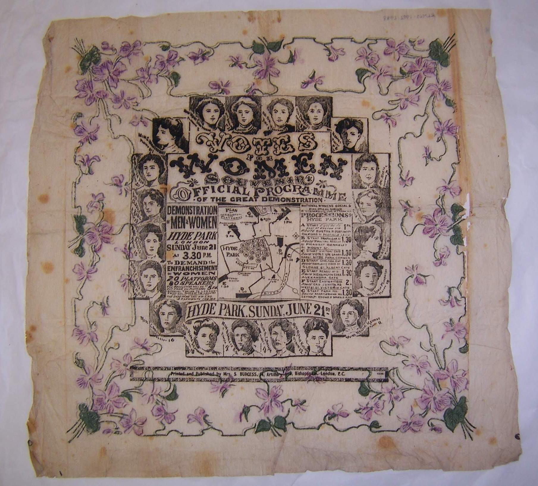 A square, aged fabric napkin with black printed text and portraits in the centre, bordered by purple flowers and green leaves. The centre features a map and decorative “Votes for Women” text. The fabric is lightly stained and worn.