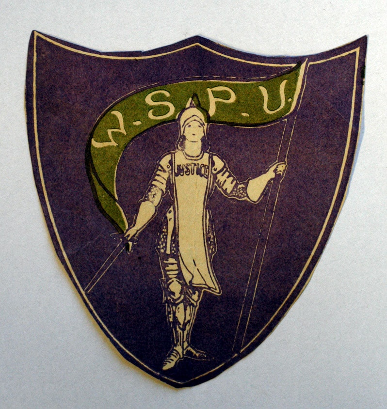 Shield-shaped paper emblem of W.S.P.U. Suffragettes using the colours green (for hope), purple (for dignity) and silver or white for purity.