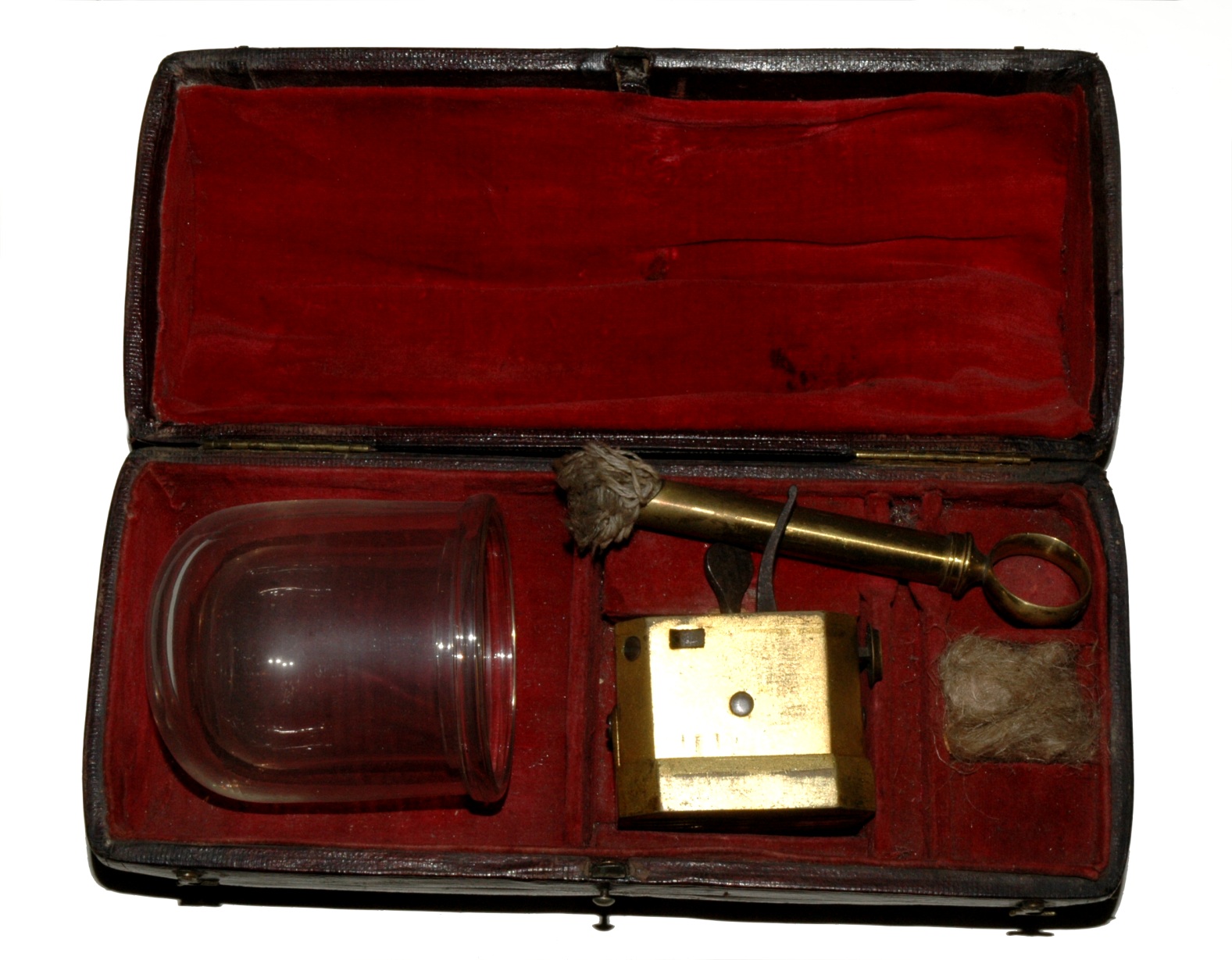 A vintage medical cupping set in an open red velvet-lined case. It includes a glass cup, brass tool, and hemp fibers.