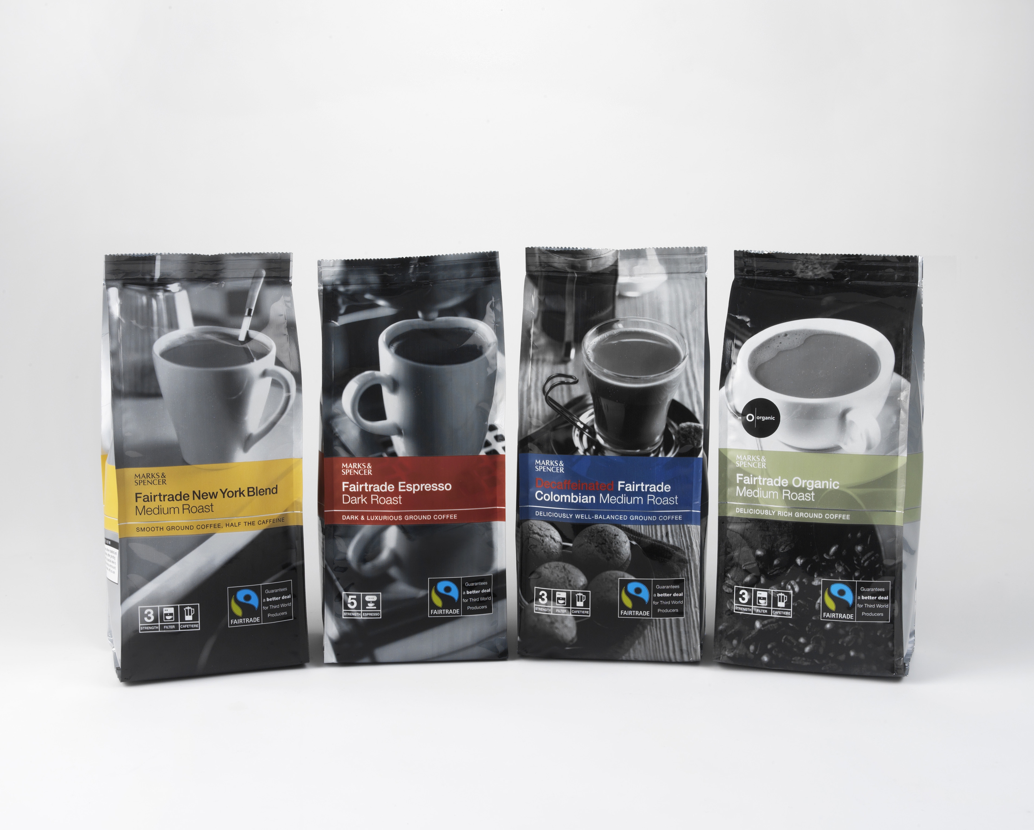 Four M&S coffee bags stand in a row, each with a black-and-white coffee cup image. Labels indicate Fairtrade blends: New York, Espresso Dark, Colombian, and Organic Medium Roasts.