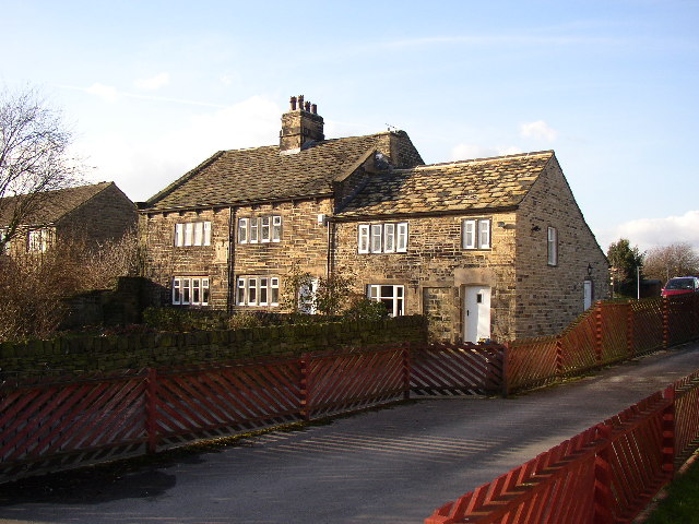A traditional weaver's cottage made of stone with a tiled roof and multiple small windows. The building is surrounded by a low stone wall and wooden fencing, situated beside a narrow road. The sky is clear and blue.