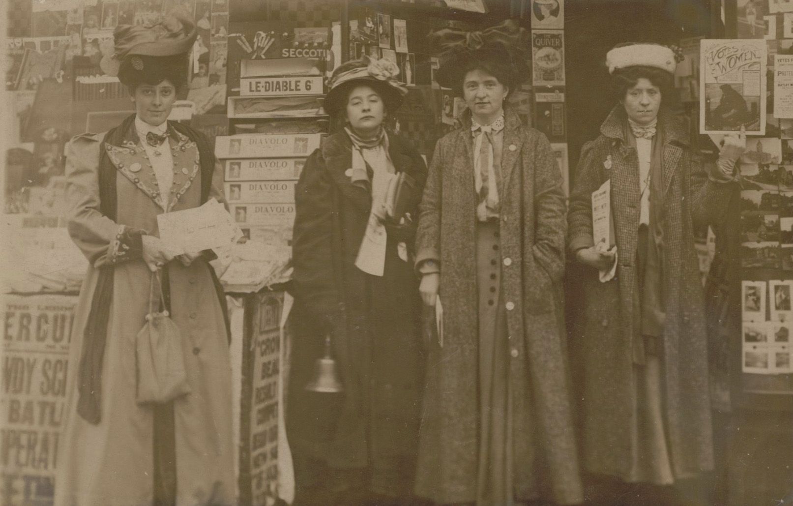 Four women dressed in early 20th-century clothing stand in a row outdoors. They wear long coats and hats, and one holds a small bell. Behind them is a wall covered in posters and advertisements. The image is in sepia tone.