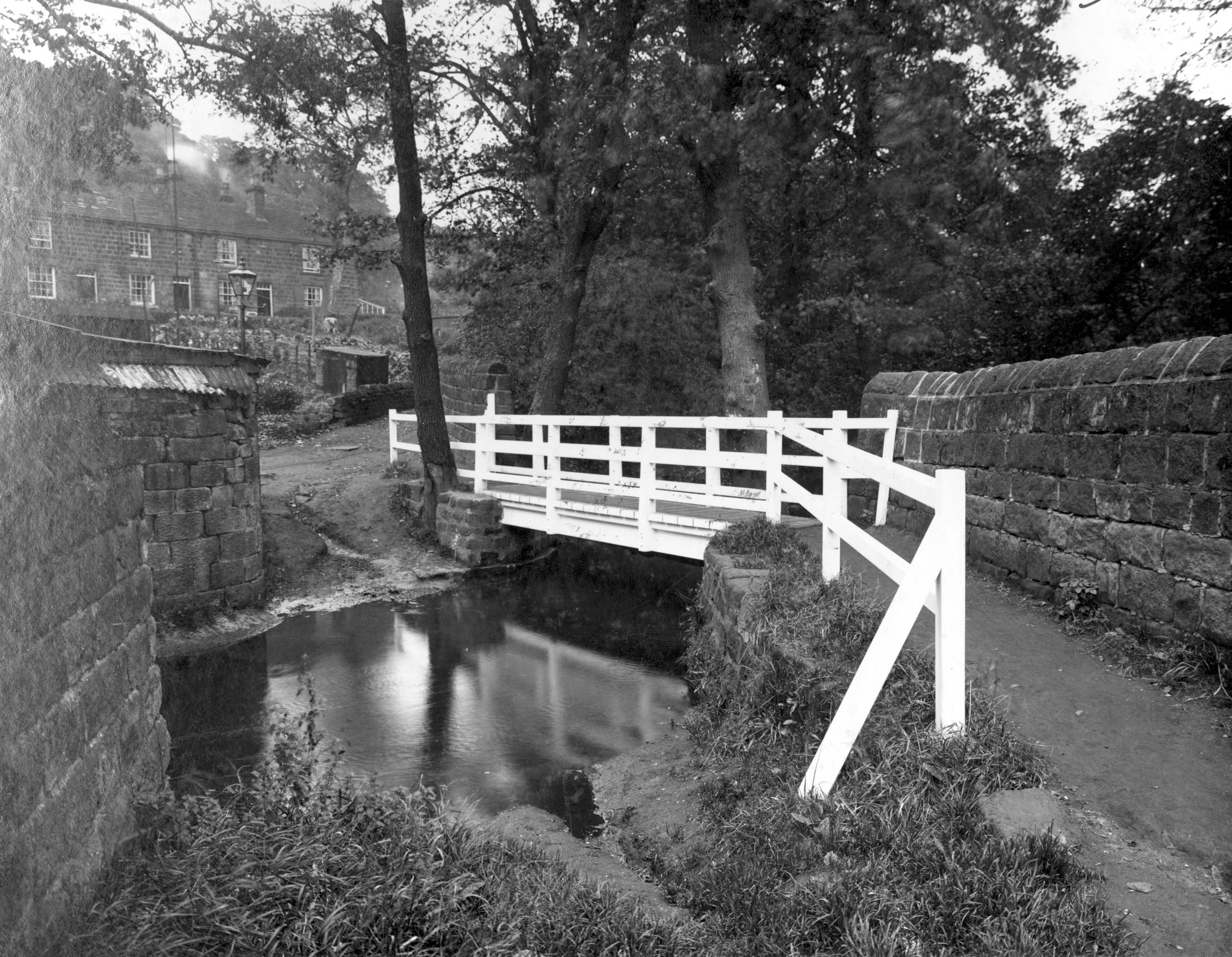 Black and white photograph showing a white wooden bridge crossing a small beck. In the background is Hustler’s row, a row of stone terraced cottages.