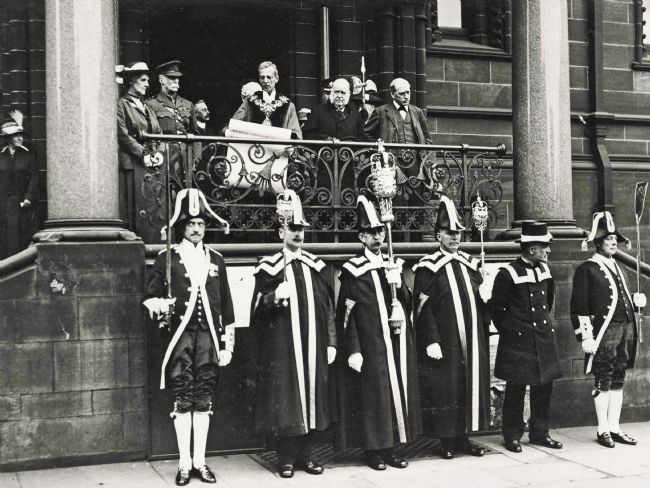 A line of men standing outside the town hall , dressed in robes of office. On the steps the proclamation is being read , maybe by the mayor.