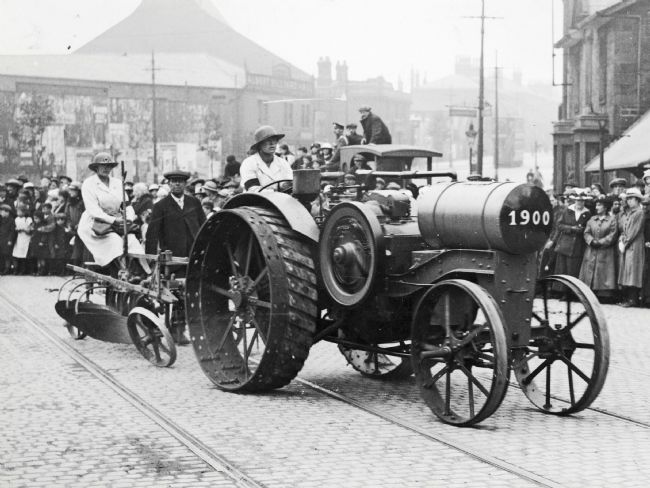WW1 Women's Land Army Driver on a Farm Tractor with a Plough driving through the streets of a town.