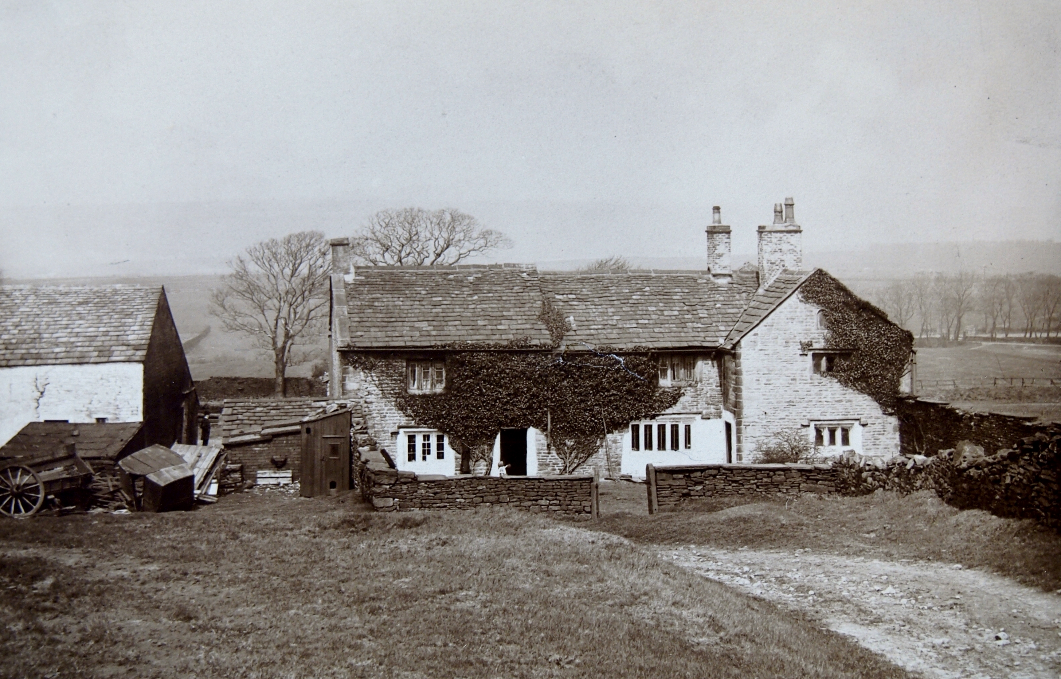 Black and white photograph showing an old farm house with plants growing up the walls of the house. It is stone built and surrounded by a stone wall. In the background and trees and fields.