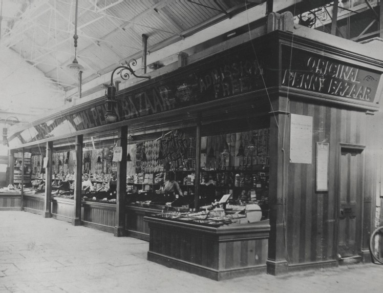 A black and white photograph of a vintage indoor market stall titled "Original Penny Bazaar." Products line the shelves, with a shopkeeper visible.