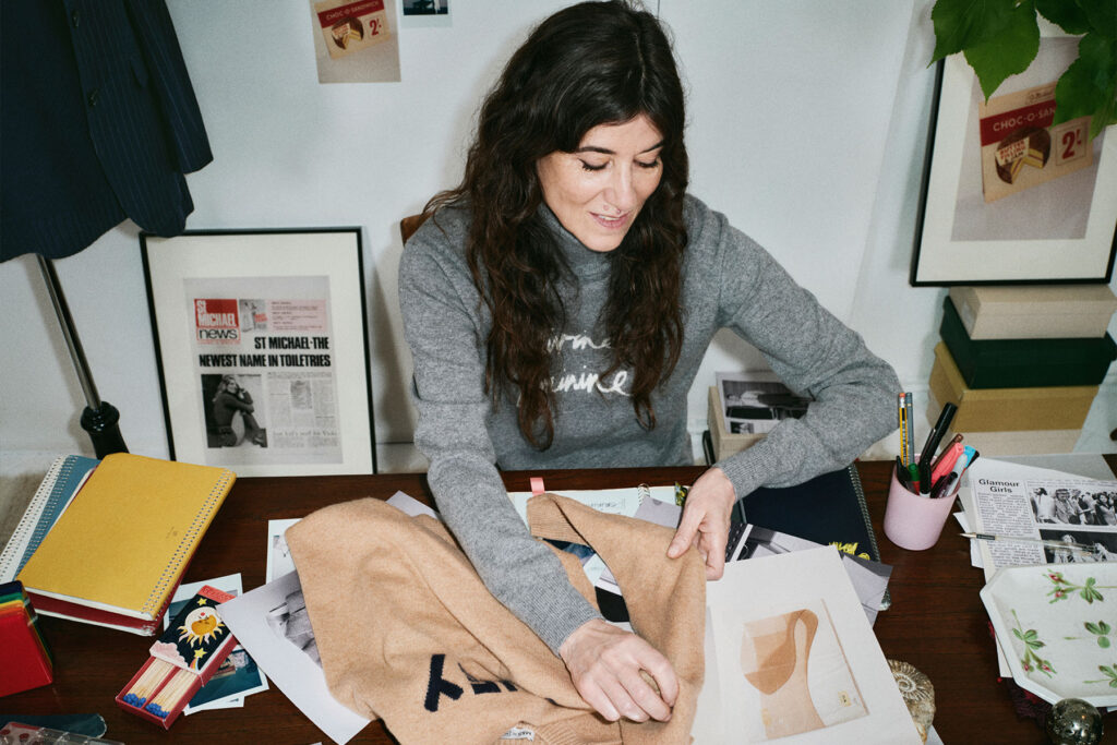 Bella Freud sitting at a desk examines a camel-coloured garment alongside design sketches and notebooks.