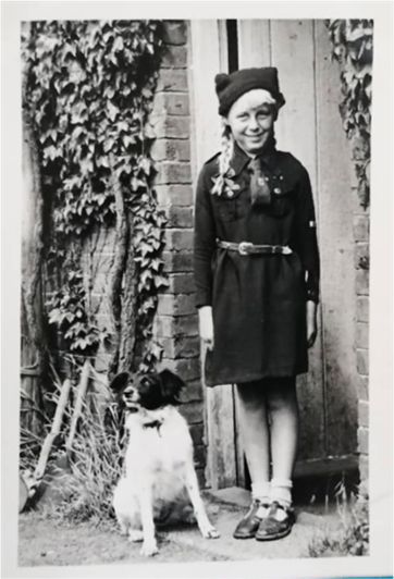 Black and white photograph showing a young girl on a doorstep. She is wearing a Brownie uniform of a long sleeved dress with a tie, belt and what looks like a knitted hat.