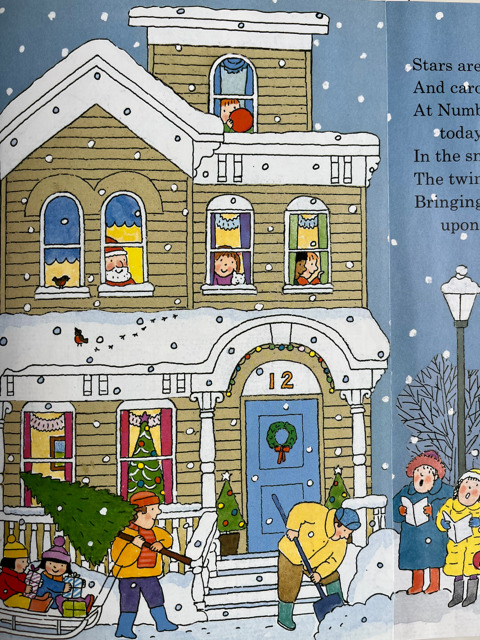An illustration from a children's book showing a festive winter scene outside a three-story house numbered 12, decorated for Christmas. Snow is falling, and children and adults are engaged in various activities. A man carries a Christmas tree while a child in a sled is pulled behind him. Another man is shoveling snow on the sidewalk. Several people are seen inside the house, looking out from the windows, including children, adults, and a cat. A Christmas wreath hangs on the front door, and lights and garlands decorate the exterior. A lamppost and two women in winter clothing stand on the right side. The edge of a poem or text is partially visible on the right-hand page.