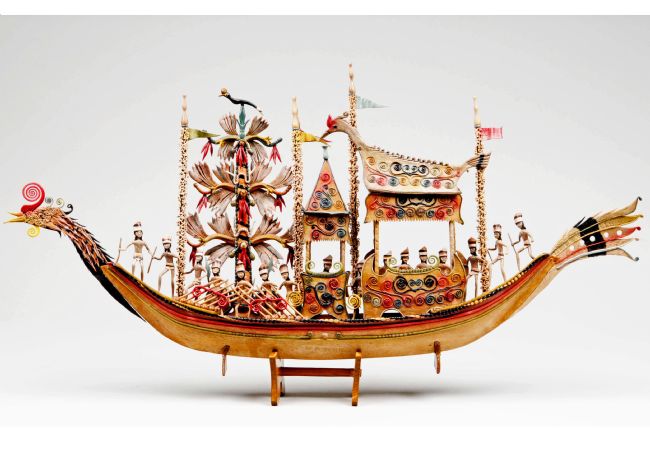 Model boat in the shape of a bird.  On board are many figures and three structures, two of which are canopies.  It is a colourful model, with yellow, red and blue the most prominent colours.