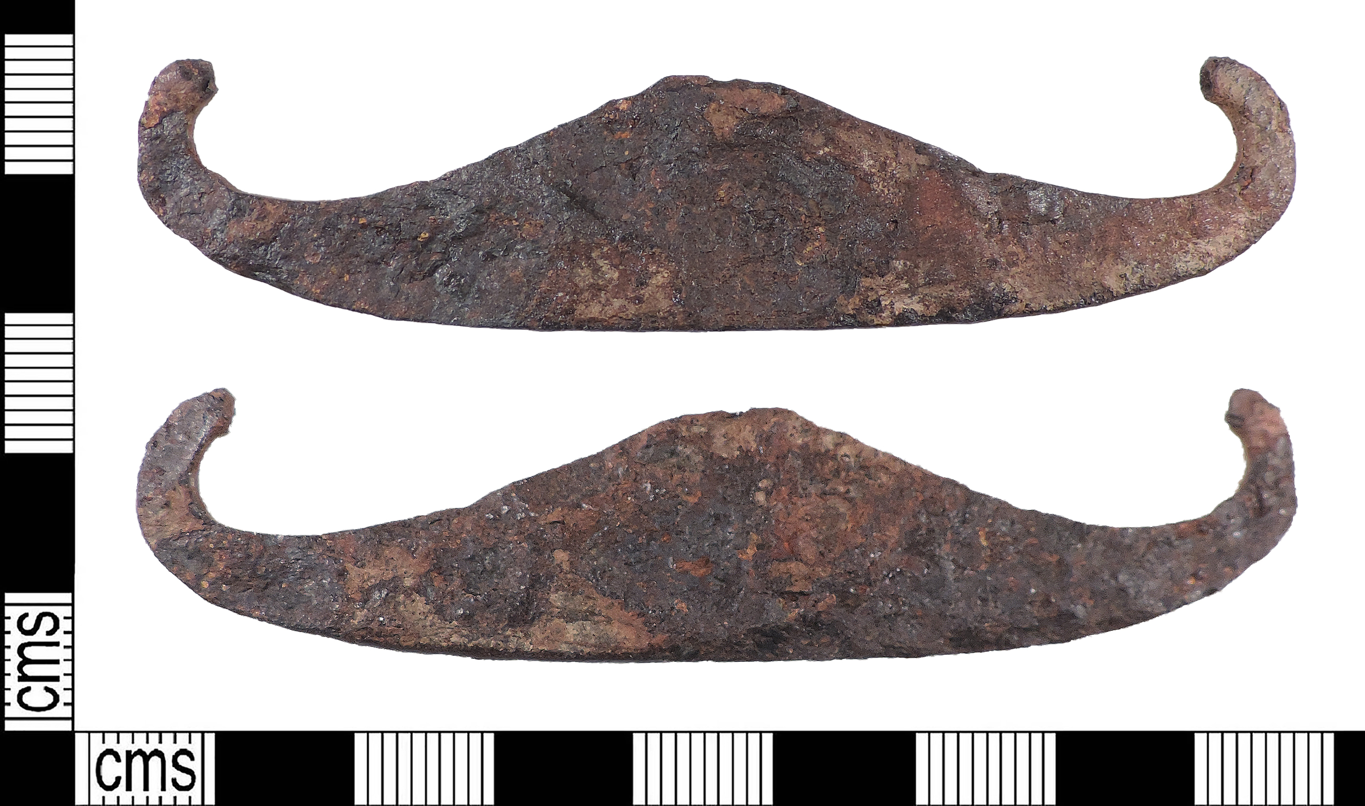 Photograph of an Early Medieval iron strike-a-light, heavily corroded with a symmetrical, moustache-like shape and upturned hooked terminals. The object is shown from both sides against a white background with a black and white centimetre scale for reference, showing it to be around 8cm long.