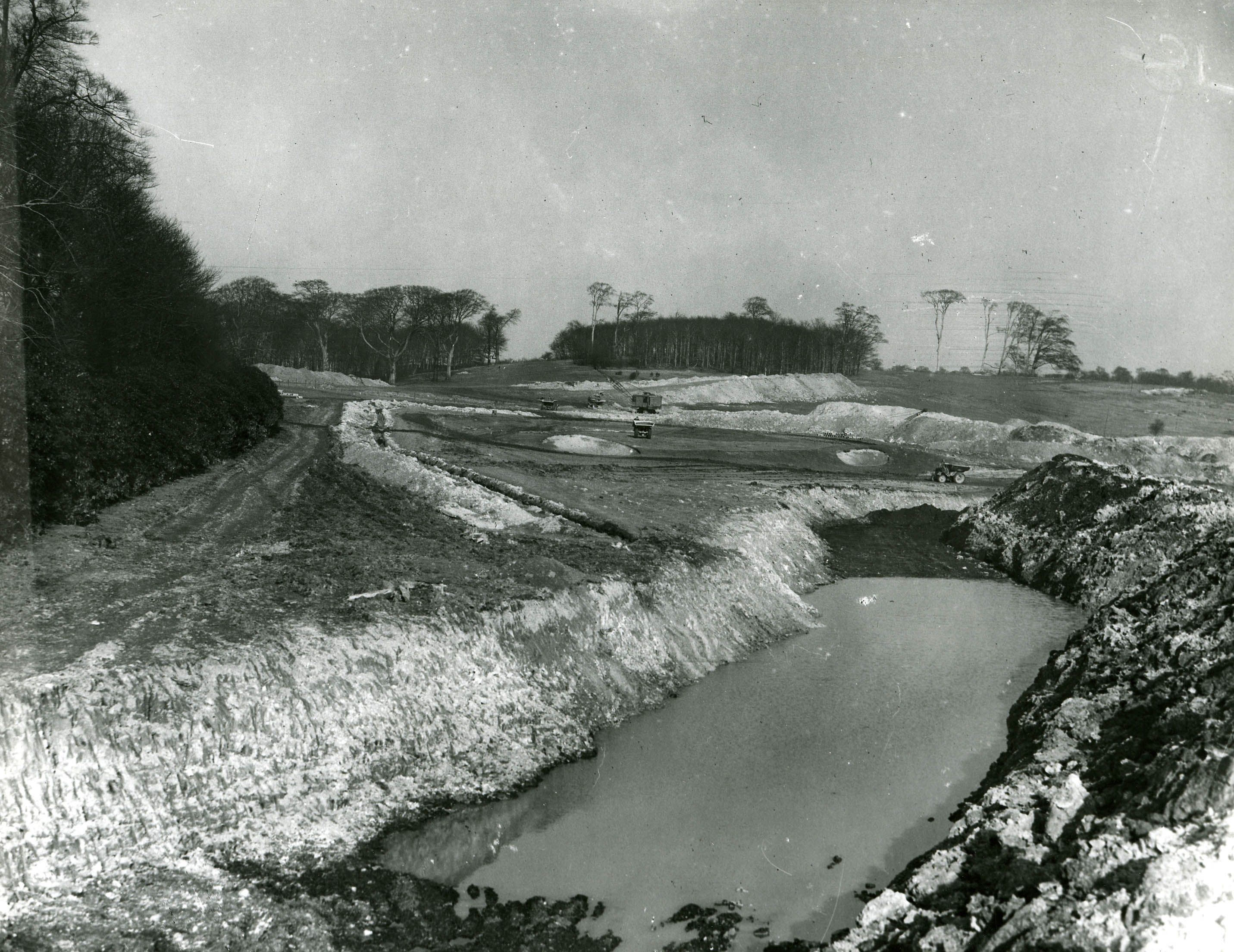 Black and white photograph showing water filled pits in a scarred landscape with trees in the distance