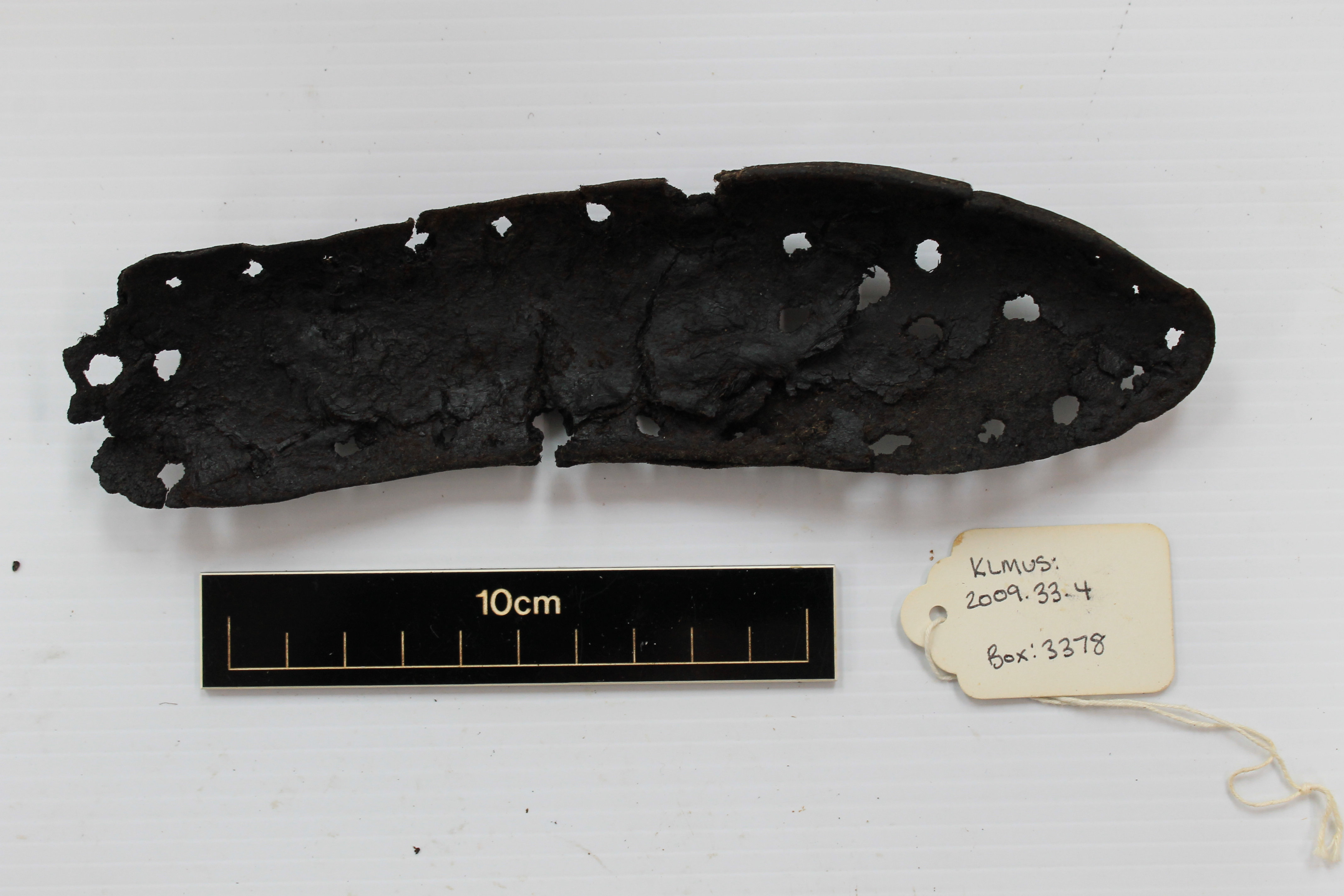 A colour photograph of a deteriorated Roman shoe sole, dark brown to black in colour, with numerous holes and signs of wear and decay. The leather object is laid flat on a white background, with a black measurement scale beneath showing a length of approximately 22 cm, leading to the belief that it was a child or woman's shoe. To the right, a cream-colored tag reads an accession number for museum cataloguing.
