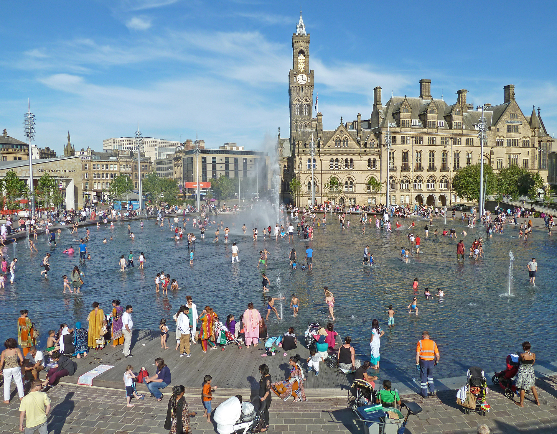 Contemporary photograph showing crowds of people on a summer day in a city centre shallow lake, with a grand building in the background