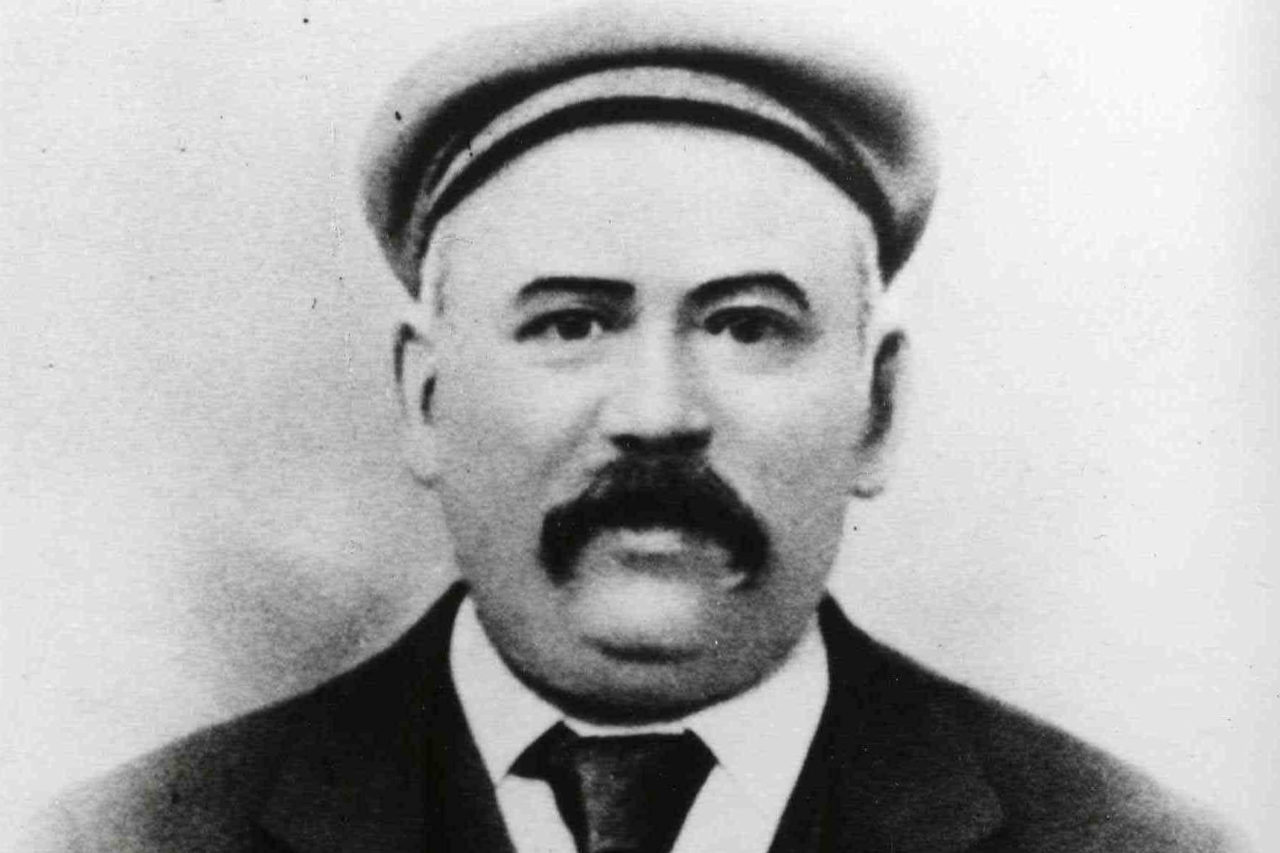 Black and white portrait photo of gentleman wearing flat cap, moustache, jacket and tie