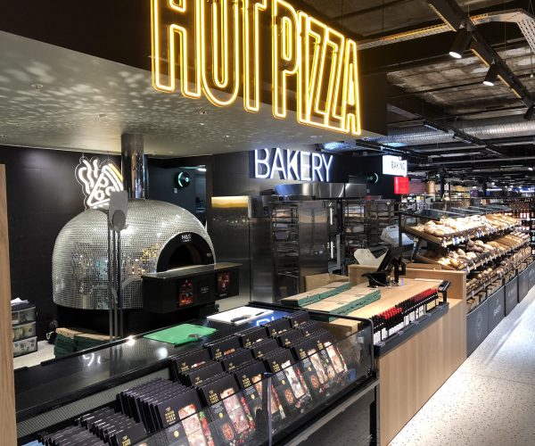 A modern bakery and pizza counter inside a Marks & Spencer store, featuring a shiny silver glitterball-themed pizza oven, illuminated "Hot Pizza" and "Bakery" signs, and a display of pastries.