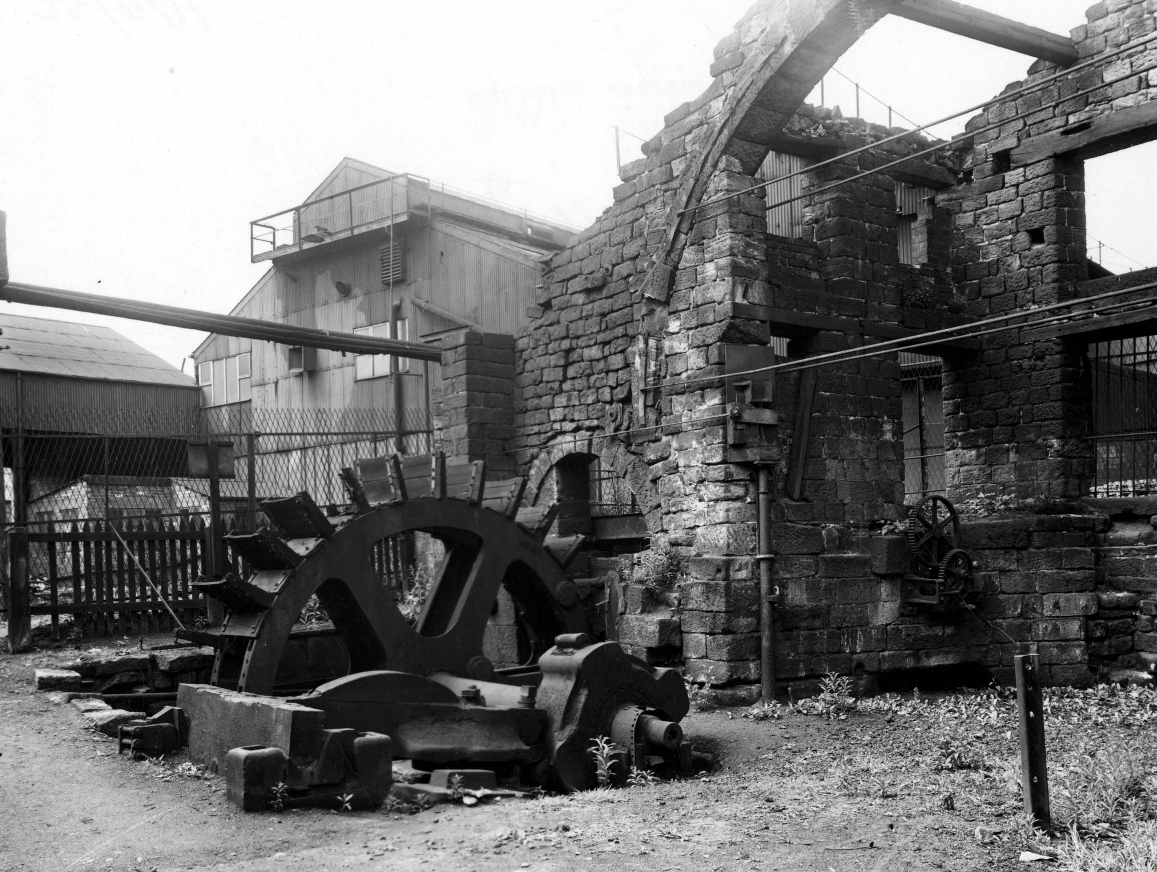 Black and white photograph of a large metal wheel, that drives a hammer